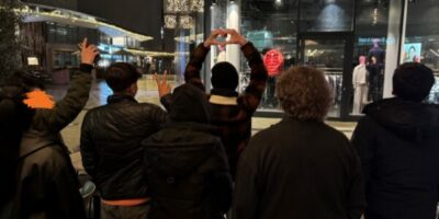 Eine Gruppe von vier Personen steht mit dem Rücken zur Kamera vor einem beleuchteten Schaufenster in einer nächtlichen Stadt. Eine Person formt mit den Händen ein Herz, während eine andere das Peace-Zeichen zeigt. Im Hintergrund sind festliche Lichter und Dekorationen zu sehen, die eine gemütliche Atmosphäre schaffen.