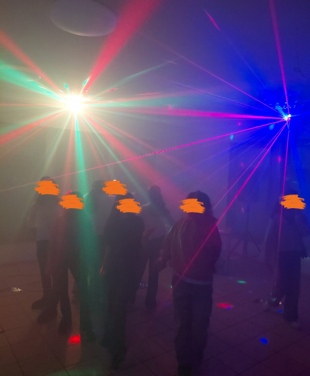 Eine Gruppe von Menschen tanzt in einem Raum mit bunten Lichteffekten und Lasern, die eine lebendige und dynamische Atmosphäre schaffen. Der Raum ist leicht neblig, wodurch die Lichtstrahlen besonders hervorgehoben werden. Die Szene vermittelt das Gefühl einer ausgelassenen Party oder Feier.