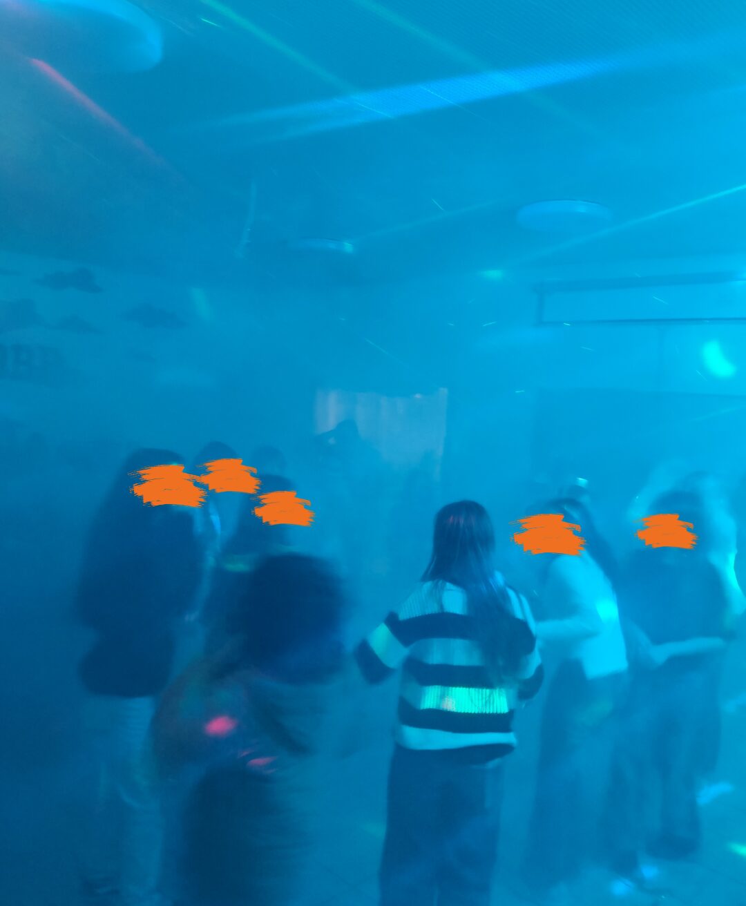 Eine Gruppe von Menschen tanzt in einem Raum mit blauer Beleuchtung und Nebeleffekten, die eine stimmungsvolle Atmosphäre schaffen. Bunte Lichtstrahlen und Reflexionen verstärken den Eindruck einer Party oder Feier. Die Personen bewegen sich ausgelassen und scheinen die Musik zu genießen.