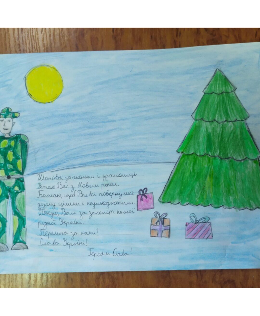Ein handgezeichnetes Bild zeigt eine winterliche Szene mit einem großen grünen Tannenbaum, mehreren bunt verpackten Geschenken und einer gelben Sonne am Himmel. Neben dem Baum steht eine Figur in grünem Tarnmuster. Darunter befindet sich ein handgeschriebener Text in ukrainischer Sprache, der Neujahrswünsche und Dankbarkeit ausdrückt.