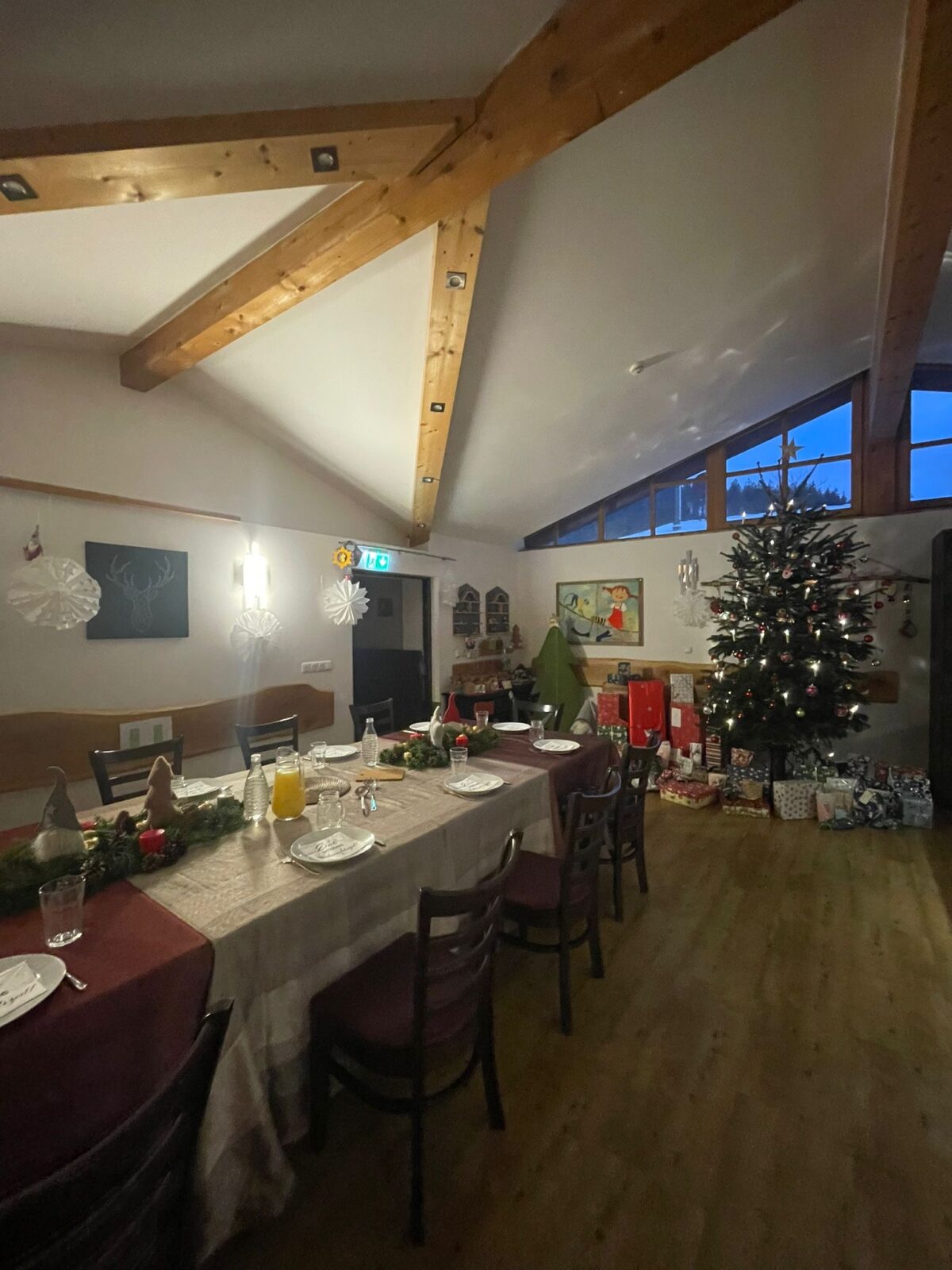 Ein festlich dekorierter Raum mit einem großen Esstisch, der für ein Weihnachtsessen gedeckt ist. Im Hintergrund steht ein geschmückter Weihnachtsbaum, umgeben von Geschenken. Die gemütliche Atmosphäre wird durch warme Beleuchtung und weihnachtliche Dekorationen unterstrichen.