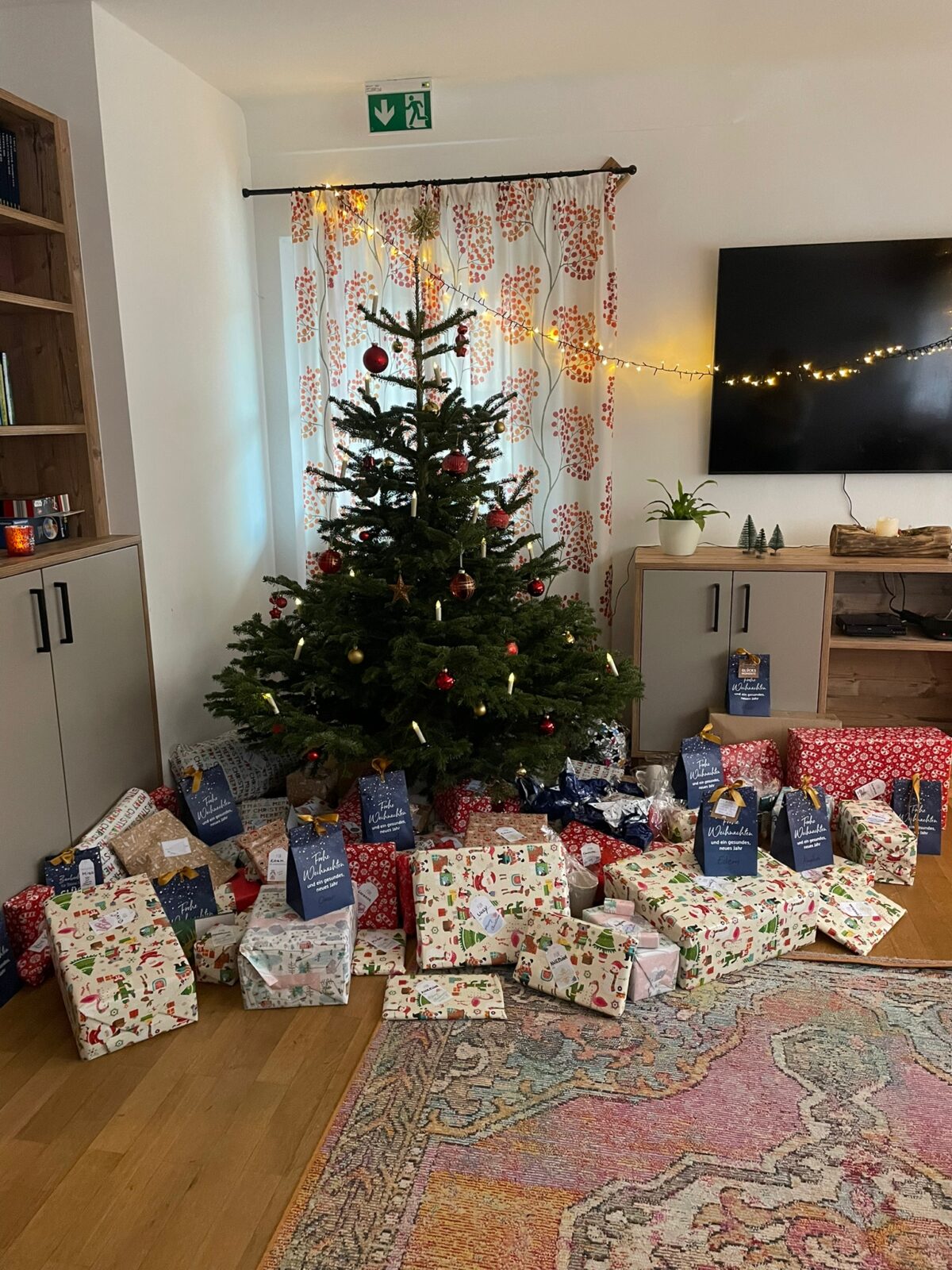 Ein geschmückter Weihnachtsbaum mit roten und goldenen Kugeln sowie Kerzen steht in einem gemütlichen Wohnzimmer. Darunter befinden sich zahlreiche bunt verpackte Geschenke, die für die Bescherung bereitliegen. Im Hintergrund sind eine dekorative Lichterkette und Vorhänge mit farbenfrohem Muster zu sehen.