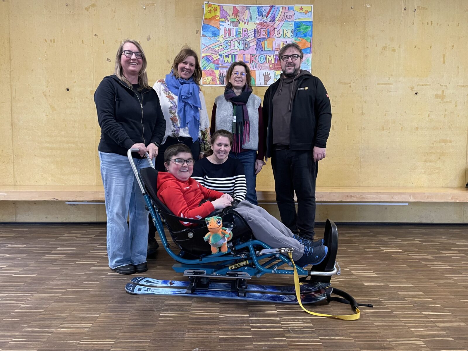 Eine Gruppe von Personen steht in einem Raum mit Holzfußboden und einer gelben Wand. Im Vordergrund sitzt eine Person in einem speziellen Skigerät, das mit einem bunten Stofftier dekoriert ist. Im Hintergrund ist ein farbenfrohes Plakat mit der Aufschrift "Hier bei uns sind alle willkommen" zu sehen.