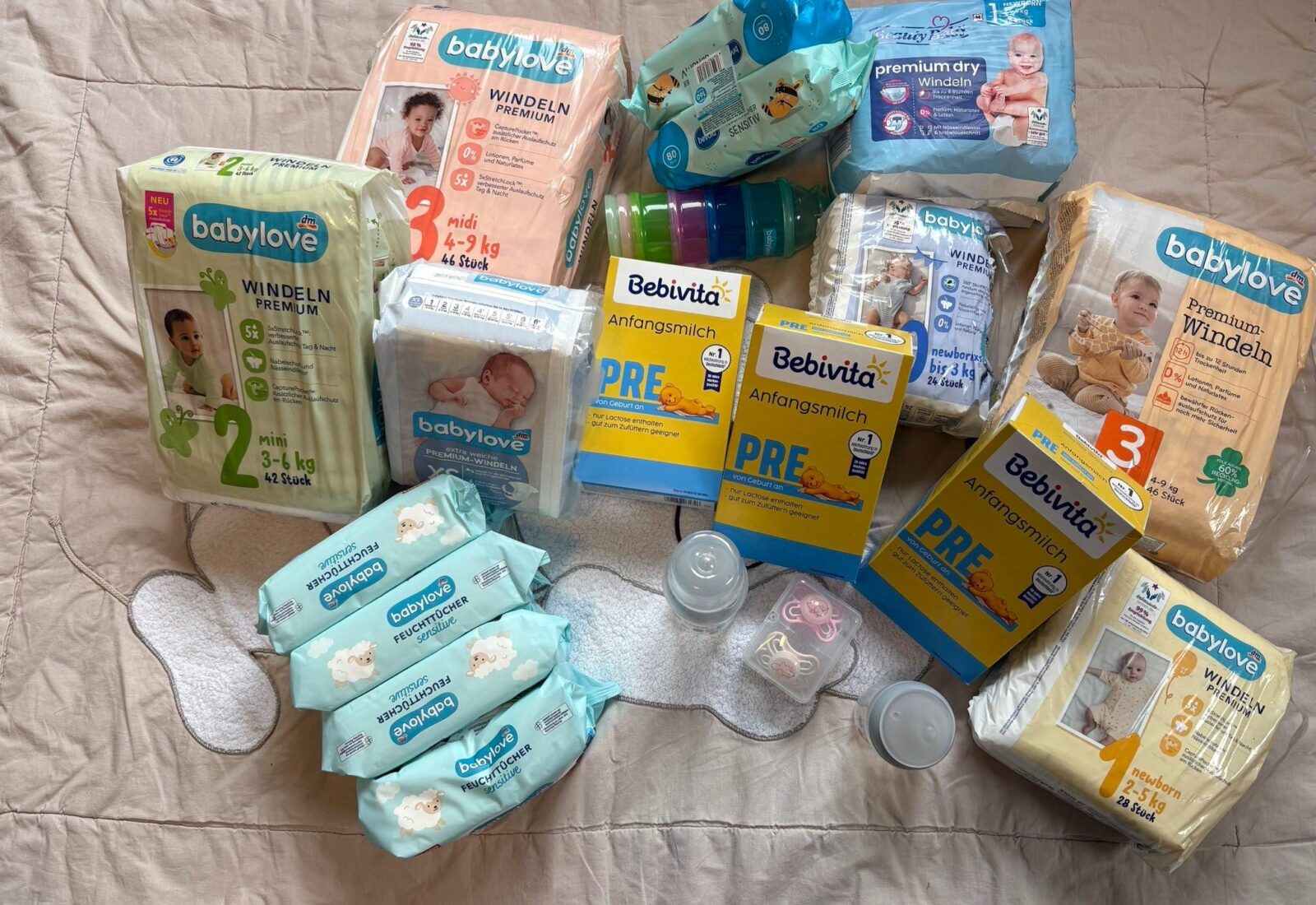 Ein Arrangement von Babyprodukten, darunter Windeln, Feuchttücher, Anfangsmilch und Babyflaschen. Die Artikel sind sorgfältig auf einer Decke ausgelegt und umfassen verschiedene Marken wie Babylove und Bebivita. Zusätzlich sind Schnuller und ein Behälter für Babynahrung sichtbar.