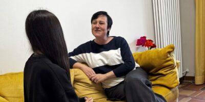 Zwei Personen sitzen auf einem gelben Sofa in einem gemütlichen Raum mit roten Teppichakzenten. Eine Person trägt eine schwarze Kleidung und graue Jogginghose, während die andere lässig gekleidet ist. Im Hintergrund sind eine Heizung, gelbe Vorhänge und eine Vase mit roten Blumen zu sehen.