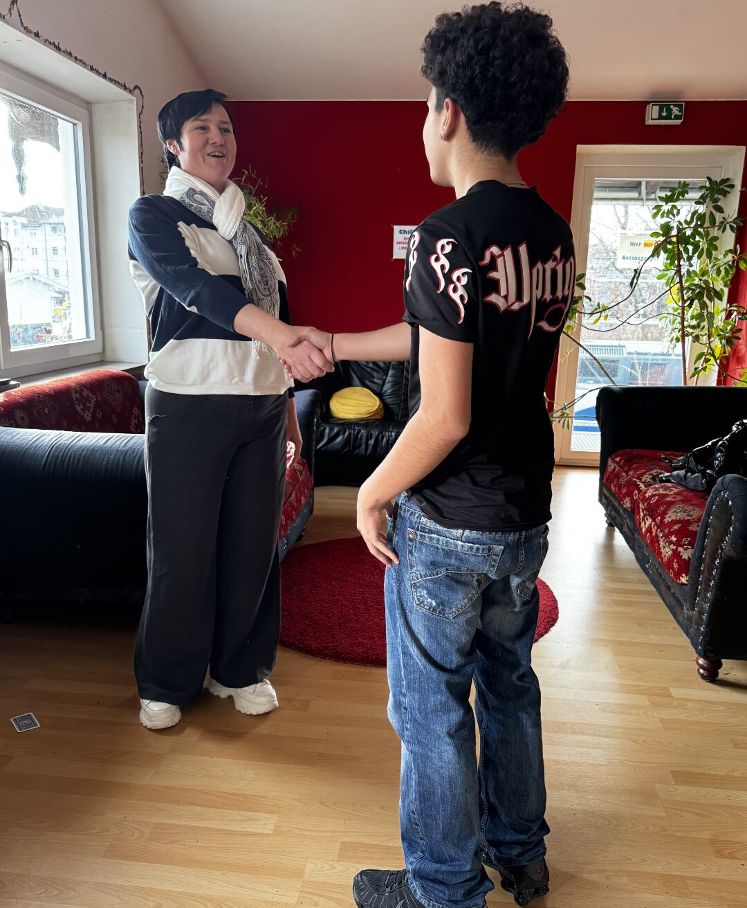 Zwei Personen stehen in einem gemütlich eingerichteten Raum mit roten Wänden und Holzboden und geben sich die Hand. Im Hintergrund sind ein Sofa, Pflanzen und ein Fenster zu sehen, das Tageslicht in den Raum lässt. Die Szene wirkt freundlich und einladend.