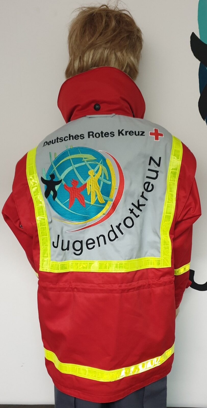 Rückansicht einer roten Einsatzjacke des Jugendrotkreuzes mit reflektierenden gelben Streifen. Auf der grauen Rückenfläche ist das Logo des Deutschen Roten Kreuzes mit einer stilisierten Weltkugel und drei farbigen Figuren zu sehen. Darüber steht der Schriftzug "Deutsches Rotes Kreuz", darunter "Jugendrotkreuz".