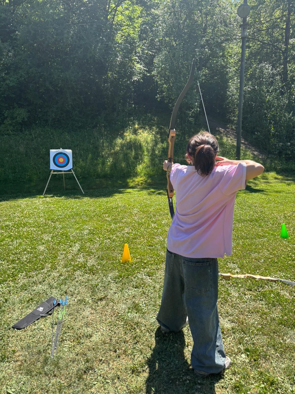 Eine Person steht auf einer grünen Wiese und zielt mit einem traditionellen Bogen auf eine Zielscheibe in der Ferne. Die Zielscheibe ist bunt und steht auf einem Ständer vor einer bewaldeten Kulisse. Neben der Person liegen Pfeile und eine Bogentasche auf dem Boden.