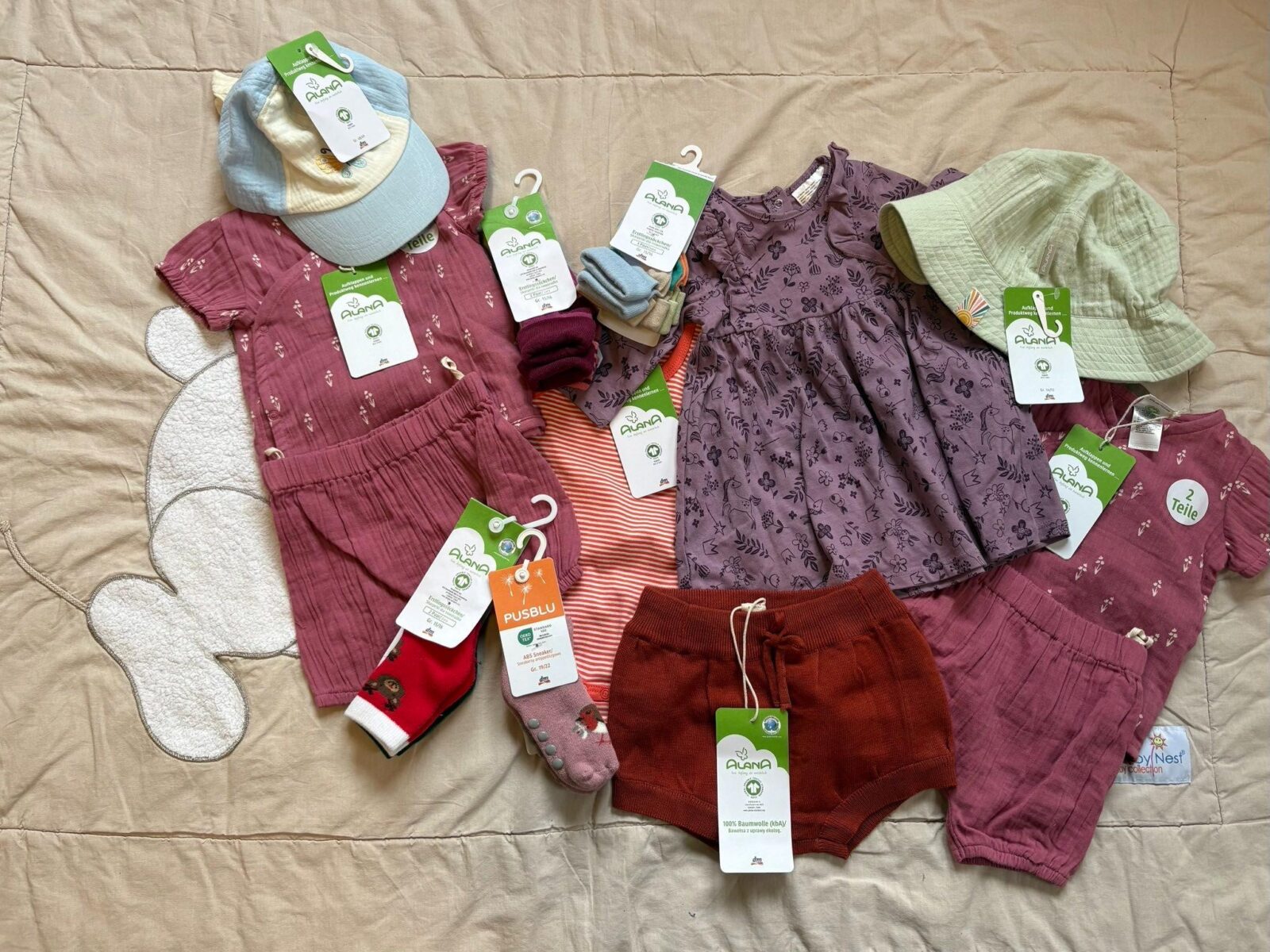 Eine Auswahl an nachhaltiger Kinderkleidung, darunter Kleidungsstücke wie Shorts, ein Kleid, Strumpfhosen und Socken, die mit Etiketten der Marke Alana versehen sind. Die Kleidung ist in warmen und natürlichen Farben wie Rot, Rosa und Lila gehalten. Zusätzlich sind auch Accessoires wie Mützen zu sehen, die auf einem beigen Untergrund arrangiert wurden.