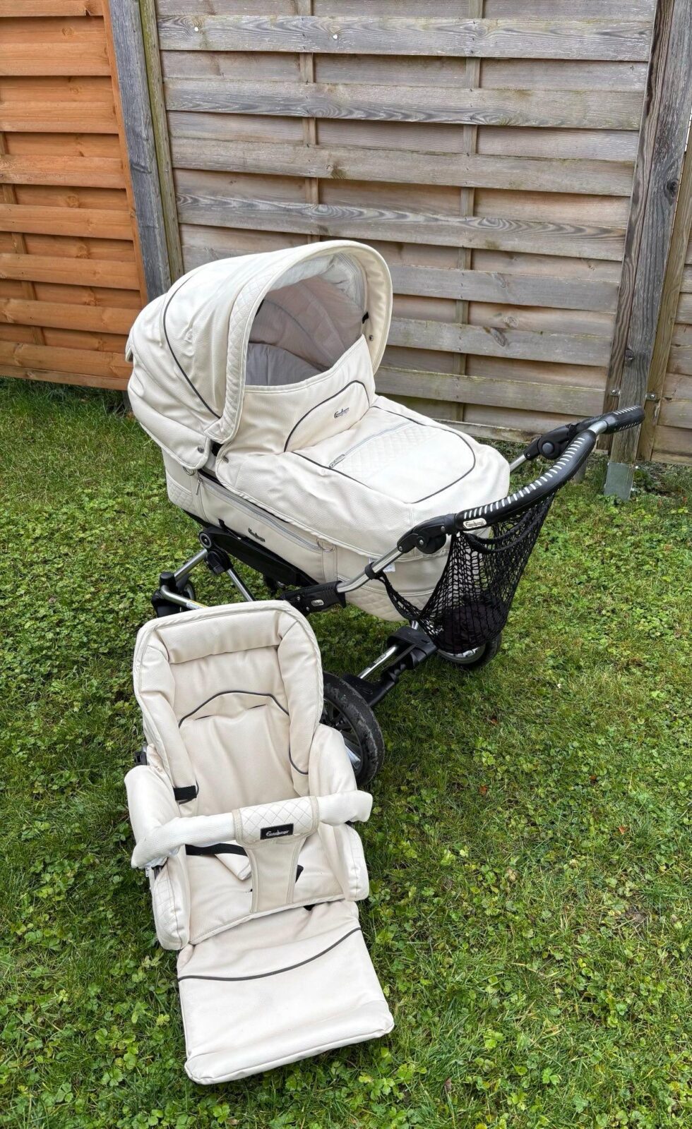 Ein beigefarbener Kinderwagen mit einem abnehmbaren Sitzaufsatz steht auf einer grünen Rasenfläche. Der Kinderwagen verfügt über ein großes Verdeck und einen praktischen Netzkorb für zusätzlichen Stauraum. Im Hintergrund ist ein Holzzaun sowie eine Holzwand zu sehen.