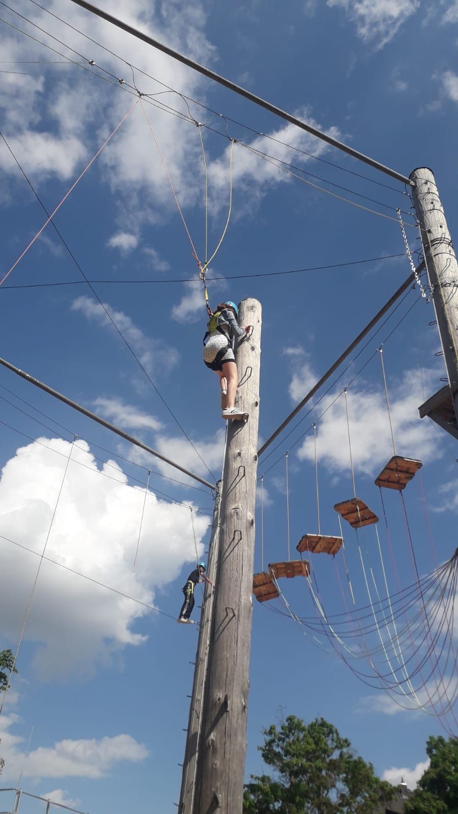 Zwei Personen klettern an einem Hochseilgarten unter freiem Himmel. Eine Person erklimmt einen hohen Holzpfahl, während die andere sich an einem Seil entlang bewegt. Der Himmel ist klar und blau mit vereinzelten Wolken, und die Konstruktion ist mit verschiedenen Hindernissen ausgestattet.