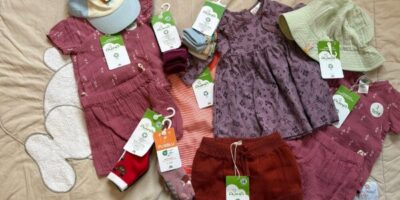 Eine Auswahl an nachhaltiger Kinderkleidung, darunter Kleidungsstücke wie Shorts, ein Kleid, Strumpfhosen und Socken, die mit Etiketten der Marke Alana versehen sind. Die Kleidung ist in warmen und natürlichen Farben wie Rot, Rosa und Lila gehalten. Zusätzlich sind auch Accessoires wie Mützen zu sehen, die auf einem beigen Untergrund arrangiert wurden.