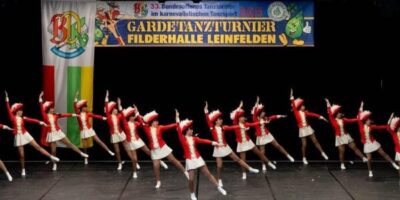 Eine Gruppe von Tänzerinnen in roten Uniformen mit weißen Röcken und passenden Hüten führt eine synchronisierte Choreografie auf einer Bühne aus. Im Hintergrund ist ein Banner zu sehen, das auf das 33. Bundesoffene Tanzturnier im karnevalistischen Tanzsport in der Filderhalle Leinfelden hinweist. Die Tanzbewegungen sind präzise und dynamisch, was die Disziplin und das Können der Tänzerinnen unterstreicht.
