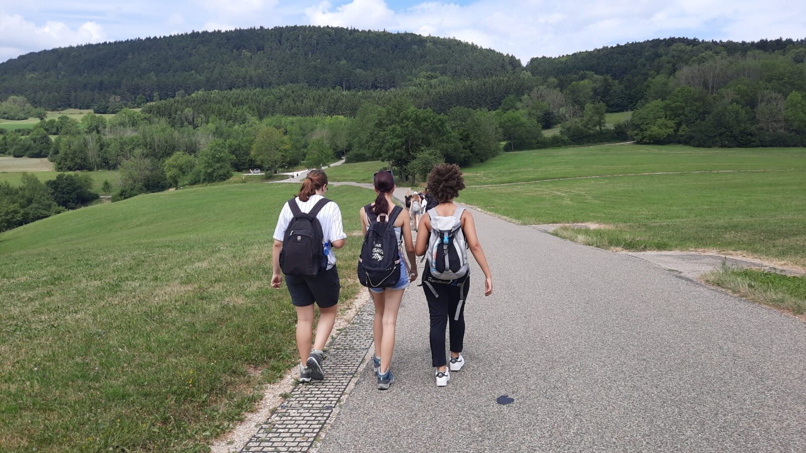 Drei junge Personen mit Rucksäcken gehen auf einem asphaltierten Weg durch eine grüne, hügelige Landschaft. Im Hintergrund sind weitere Menschen zu sehen, die ebenfalls spazieren oder wandern. Der Weg führt durch eine idyllische Umgebung mit Wiesen und einem dichten Wald im Hintergrund.