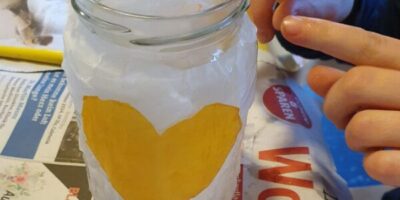 Ein Glas mit einer weißen, strukturierten Oberfläche und einem gelben Herz in der Mitte wird gebastelt. Zwei Hände sind zu sehen, die vorsichtig an der Dekoration arbeiten. Im Hintergrund liegt Zeitungspapier, das als Unterlage dient.