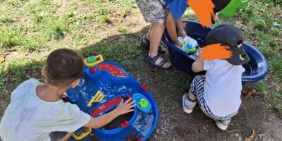 Drei Kinder spielen im Freien mit einer blauen Wasserbahn und einem blauen Wasserbecken. Sie sind in sommerlicher Kleidung und konzentrieren sich auf das Spiel mit kleinen Booten und Wasser. Der Hintergrund zeigt eine grüne Wiese und einen sonnigen Tag.
