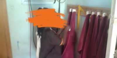 Ein Kind steht in einem Raum und beschäftigt sich mit roten Stoffen, die an Haken aufgehängt sind. Im Hintergrund sind technische Geräte an der Wand montiert, und auf dem Boden steht ein weißer Eimer. Der Raum wirkt funktional und einfach eingerichtet.