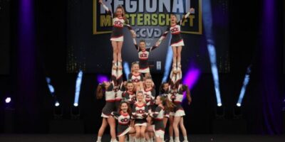 Eine Cheerleading-Gruppe führt eine beeindruckende Pyramidenformation auf einer Bühne bei der Regionalmeisterschaft des CCVD vor. Die Athletinnen und Athleten tragen einheitliche rot-weiß-schwarze Outfits mit dem Vereinslogo. Im Hintergrund ist ein beleuchtetes Banner mit dem Titel der Veranstaltung sichtbar.