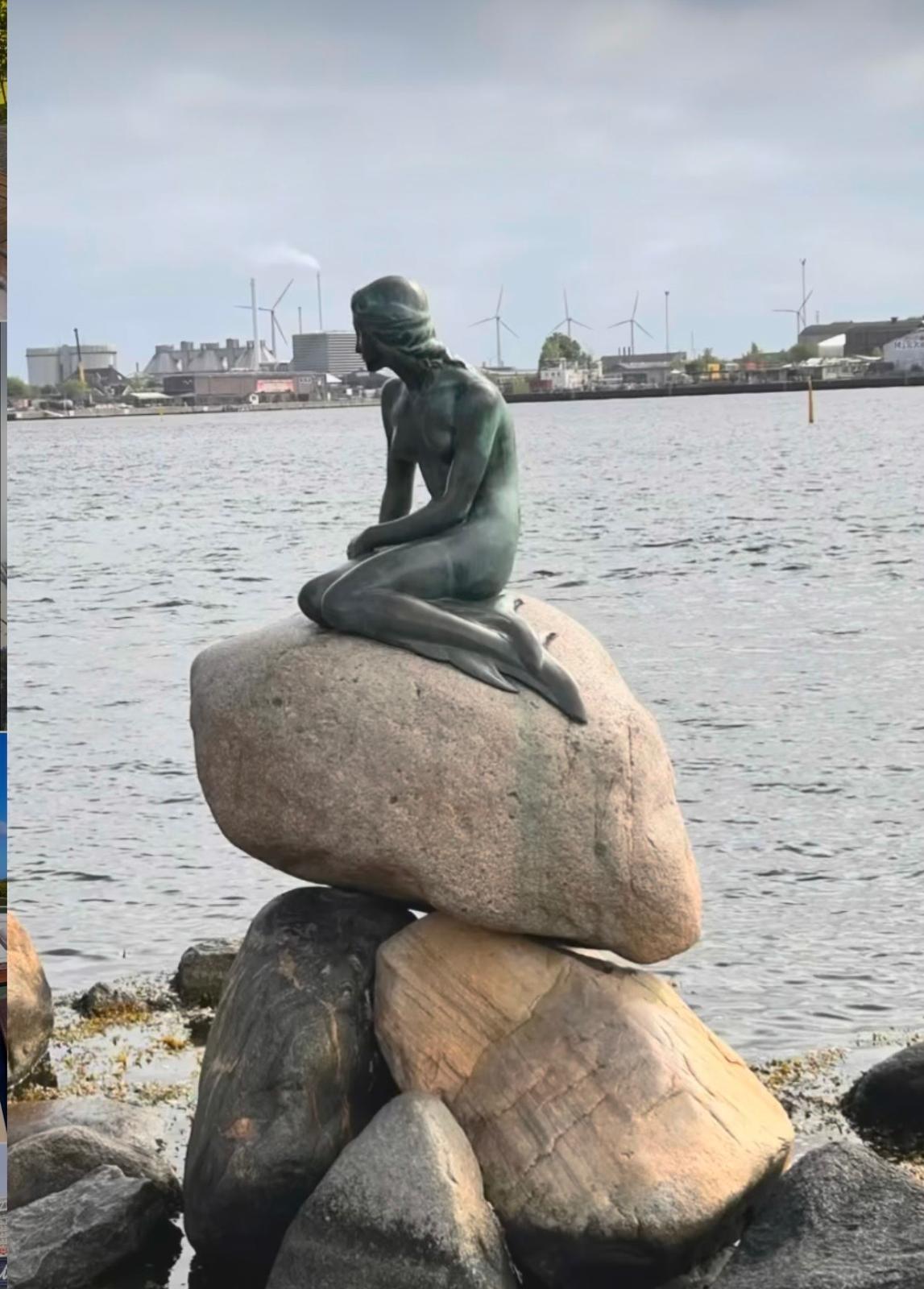 Die berühmte Statue der kleinen Meerjungfrau sitzt auf einem großen Stein am Wasser und blickt nachdenklich in die Ferne. Im Hintergrund sind Windräder und industrielle Gebäude zu sehen, die die Hafenkulisse prägen. Der Himmel ist bewölkt, was der Szene eine ruhige und melancholische Atmosphäre verleiht.