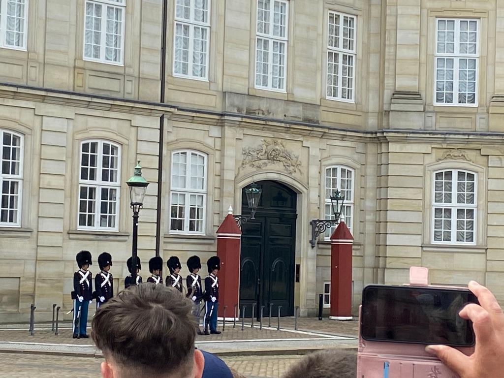 Eine historische Gebäudefassade mit großen weißen Fenstern und verzierten Steinornamenten. Im Vordergrund hält eine Person ein Smartphone, das in einer rosafarbenen Hülle steckt, und fotografiert die Szene. Neben dem Eingang des Gebäudes sind rote Säulen mit Lampen zu sehen.