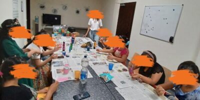 Eine Gruppe von Menschen sitzt an einem langen Tisch in einem Raum mit hellem Oberlicht. Auf dem Tisch liegen Zeitungen, Bastelmaterialien, Getränke und persönliche Gegenstände, während die Personen kreativ arbeiten. Im Hintergrund ist ein Whiteboard sowie weiteres Zubehör zu sehen.