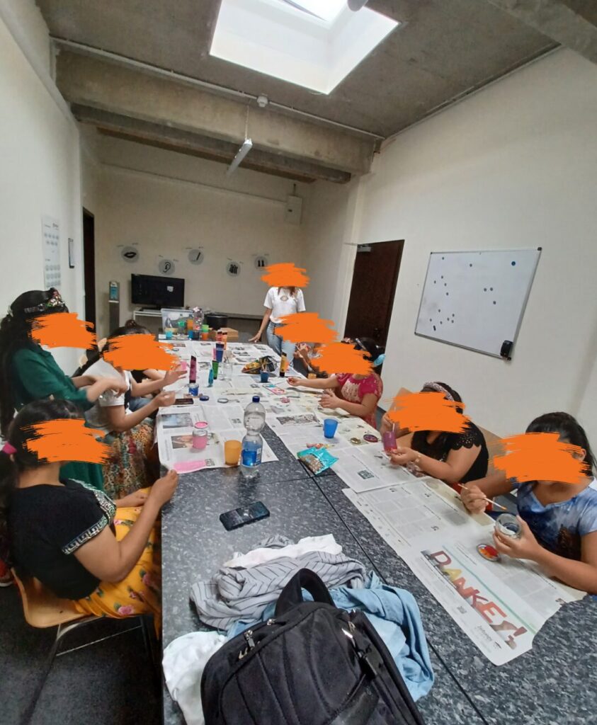 Eine Gruppe von Menschen sitzt an einem langen Tisch in einem Raum mit hellem Oberlicht. Auf dem Tisch liegen Zeitungen, Bastelmaterialien, Getränke und persönliche Gegenstände, während die Personen kreativ arbeiten. Im Hintergrund ist ein Whiteboard sowie weiteres Zubehör zu sehen.