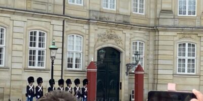 Eine historische Gebäudefassade mit großen weißen Fenstern und verzierten Steinornamenten. Im Vordergrund hält eine Person ein Smartphone, das in einer rosafarbenen Hülle steckt, und fotografiert die Szene. Neben dem Eingang des Gebäudes sind rote Säulen mit Lampen zu sehen.