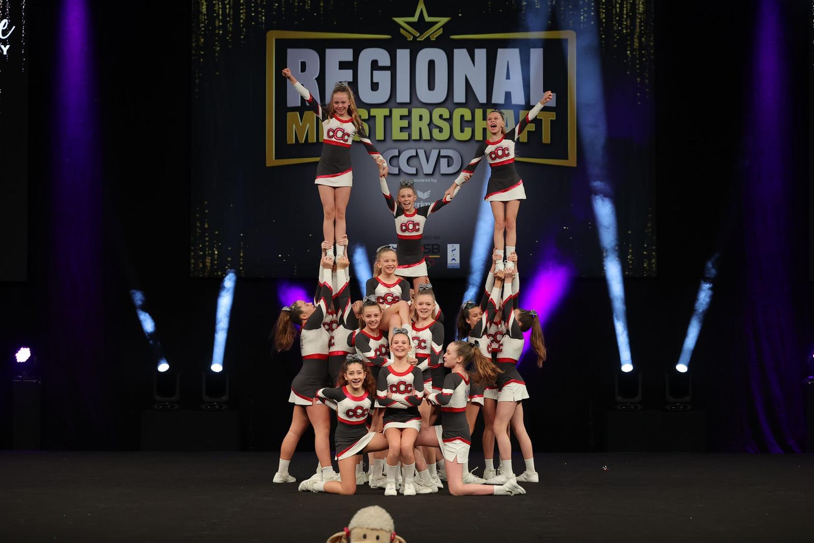 Eine Cheerleading-Gruppe führt eine beeindruckende Pyramidenformation auf einer Bühne bei der Regionalmeisterschaft des CCVD vor. Die Athletinnen und Athleten tragen einheitliche rot-weiß-schwarze Outfits mit dem Vereinslogo. Im Hintergrund ist ein beleuchtetes Banner mit dem Titel der Veranstaltung sichtbar.