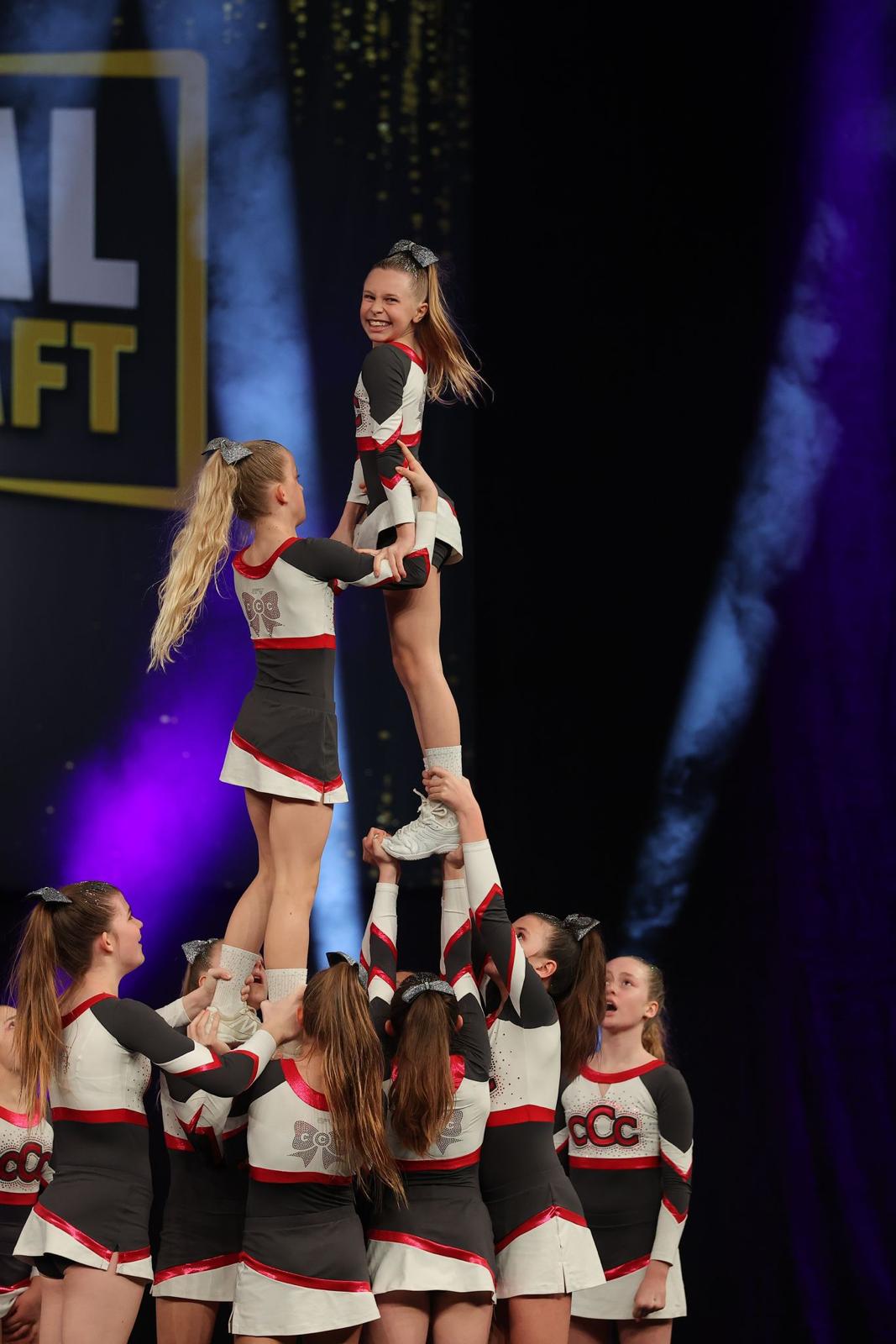 Eine Gruppe von Cheerleadern führt eine beeindruckende Hebefigur auf einer Bühne aus. Die Performerinnen tragen passende schwarz-weiß-rote Cheerleading-Outfits mit glänzenden Haarbändern. Im Hintergrund sind bunte Lichteffekte und ein Teil eines Schildes zu sehen, die die dynamische Atmosphäre des Events unterstreichen.