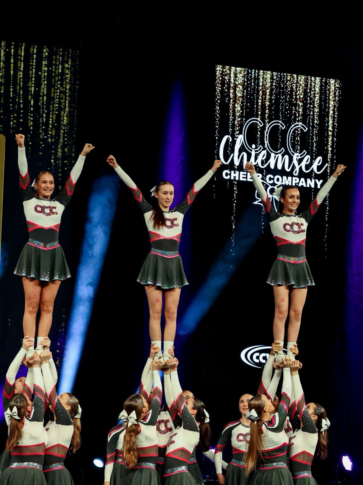 Eine Cheerleading-Gruppe führt eine beeindruckende Hebefigur auf einer Bühne aus. Die Athletinnen tragen einheitliche Outfits in Schwarz, Weiß und Rot mit glitzernden Details. Im Hintergrund sind goldene Vorhänge und das Logo der Chiemsee Cheer Company zu sehen, beleuchtet von Scheinwerfern.