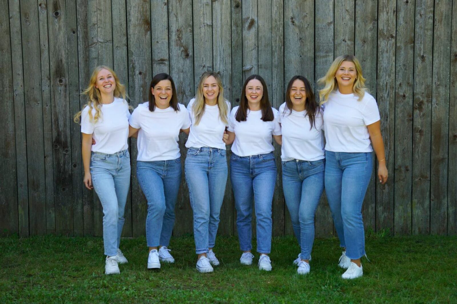 Sechs Frauen stehen nebeneinander vor einer Holzwand und tragen weiße T-Shirts und blaue Jeans. Sie haben die Arme umeinander gelegt und wirken entspannt und fröhlich. Der Hintergrund aus Holz und die grüne Wiese im Vordergrund verleihen der Szene eine natürliche und harmonische Atmosphäre.