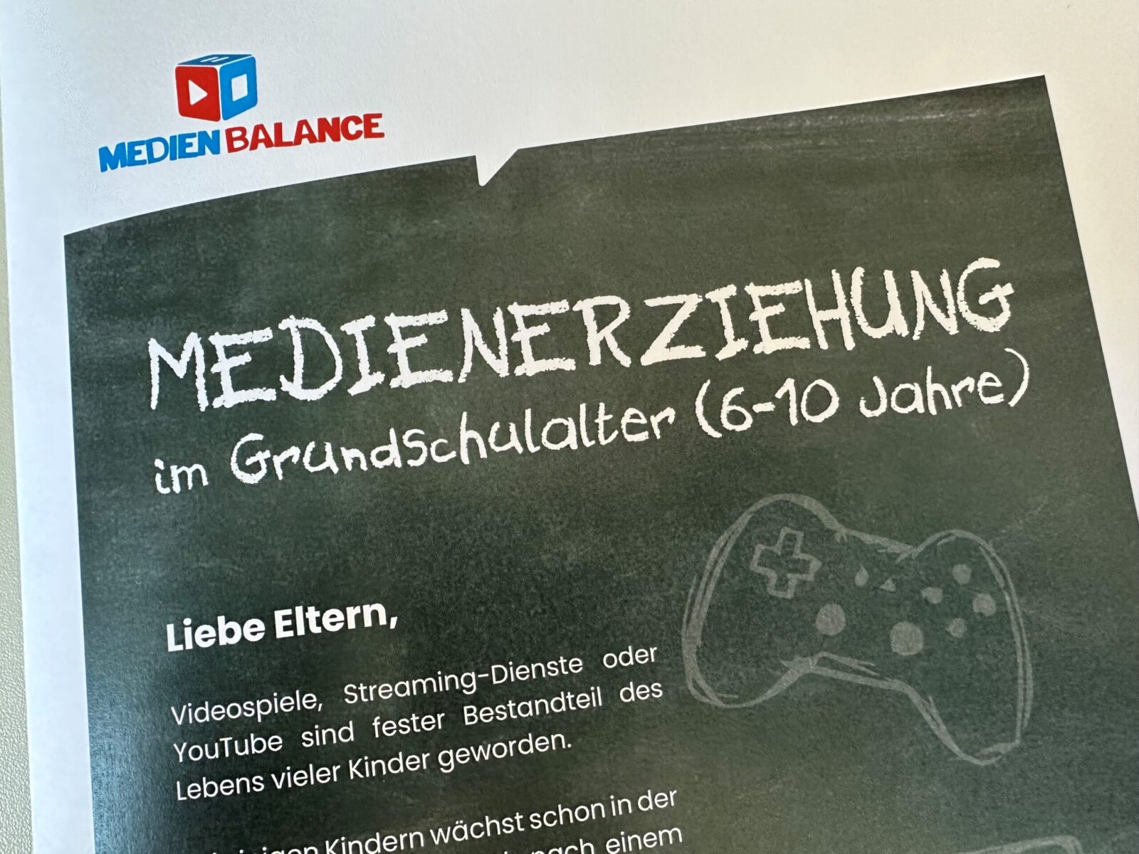 Flyer mit der Überschrift „Medienerziehung im Grundschulalter (6–10 Jahre)“, gestaltet auf einem Tafel-Hintergrund. Darunter ein gezeichneter Controller und ein Text, der sich an Eltern richtet, um über den Umgang mit Medien wie Videospielen, Streaming-Diensten und YouTube im Alltag von Kindern zu informieren. Oben links das Logo von „MedienBalance“.