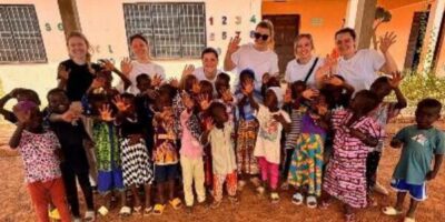 Eine Gruppe von Erwachsenen und Kindern posiert gemeinsam für ein fröhliches Gruppenfoto vor einem Schulgebäude mit orangefarbener Fassade. Die Kinder tragen bunte Kleidung und winken in die Kamera, während die Erwachsenen lächeln. Im Hintergrund sind Fenster und eine Wand mit Zahlen und Buchstaben zu sehen, die auf eine Schulumgebung hinweisen.