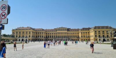Viele Menschen besuchen die Schönbrunn-Residenz in Wien, Österreich, eine weitläufige barocke Palastanlage mit prächtigen Fassaden und großen Fenstern. Die Besucher spazieren auf einem gepflasterten Platz vor dem Palast, und die Sonne scheint von einem blauen Himmel.