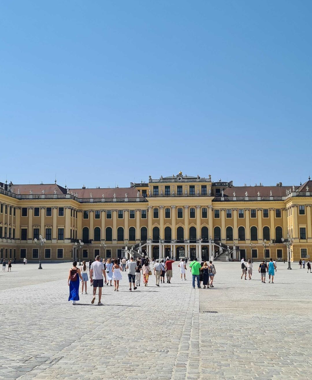 Viele Menschen besuchen die Schönbrunn-Residenz in Wien, Österreich, eine weitläufige barocke Palastanlage mit prächtigen Fassaden und großen Fenstern. Die Besucher spazieren auf einem gepflasterten Platz vor dem Palast, und die Sonne scheint von einem blauen Himmel.