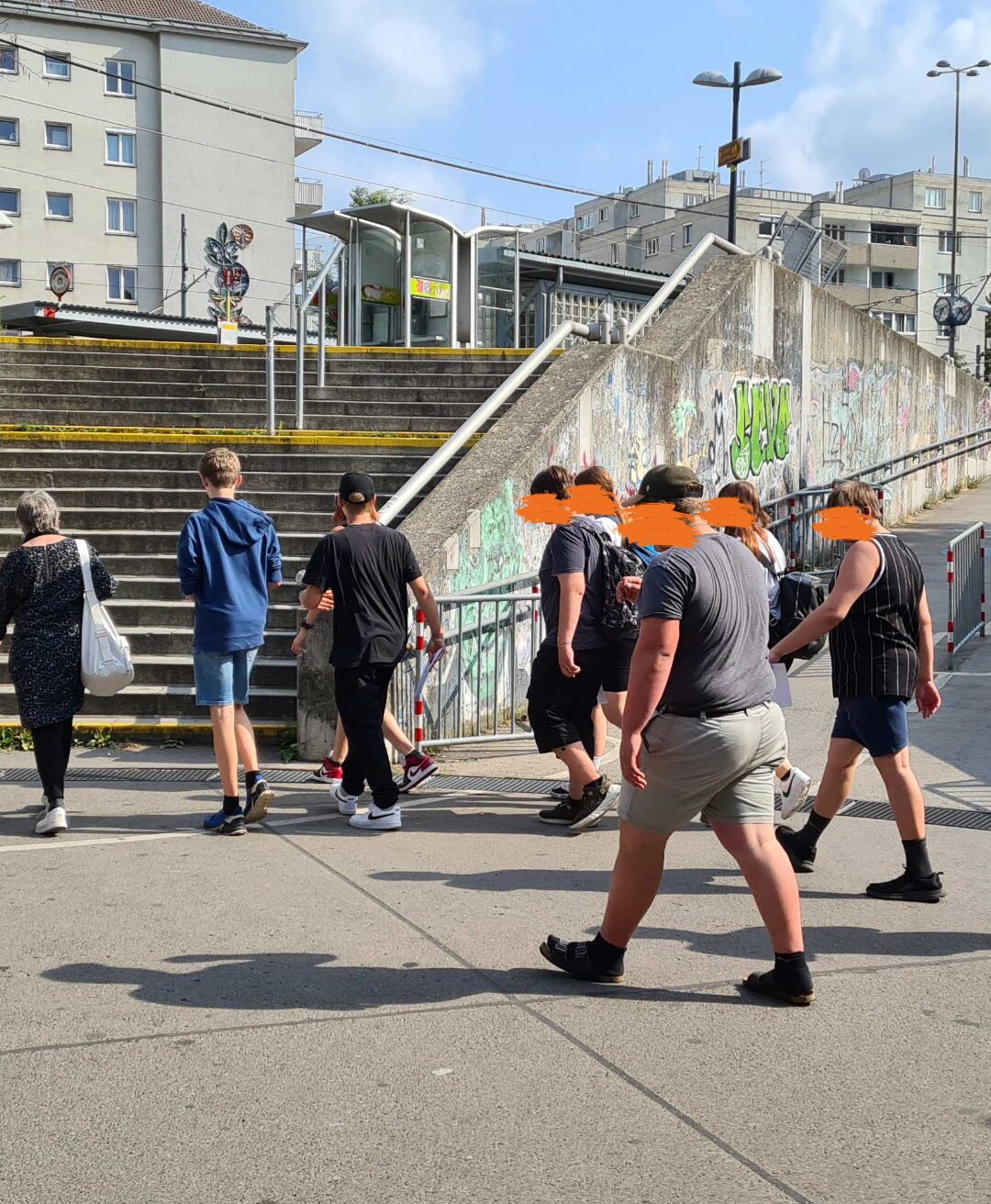 Eine Gruppe von Menschen geht über einen Gehweg, der mit Graffiti bedeckt ist. Sie gehen in Richtung einer Treppe, die zu einer Brücke führt. Ein paar Personen haben ihre Gesichter mit orangefarbenen Quadraten verdeckt.
