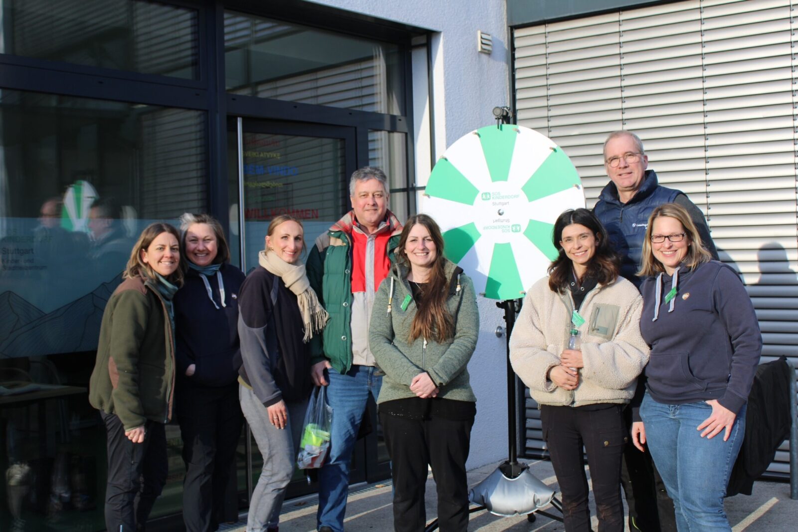 Eine Gruppe von sechs Frauen und zwei Männern stehen vor einem Gebäude mit Glasfront. Die Gruppe besteht aus vier Frauen und einem Mann. Die Frauen tragen Jacken und Schals, der Mann eine grüne Jacke. Sie alle lächeln in die Kamera.
