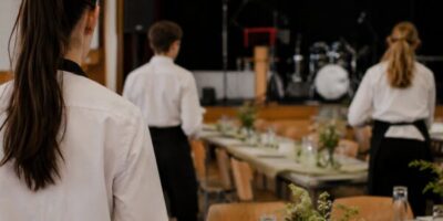 Drei Personen, gekleidet in weißen Hemden und schwarzen Schürzen, stehen in einem Saal vor einem festlich gedeckten Tisch. Der Saal ist mit Musikinstrumenten und Bühnenbeleuchtung ausgestattet, was auf eine bevorstehende Veranstaltung oder einen besonderen Anlass hinweist.