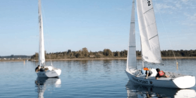 Zwei weiße Segelboote schwimmen auf dem Wasser. Auf den Segeln stehen die Buchstaben G, E, R und S. Die Besatzungsmitglieder sind nicht deutlich zu erkennen. Im Hintergrund sind Bäume und eine blaue Himmel zu sehen.