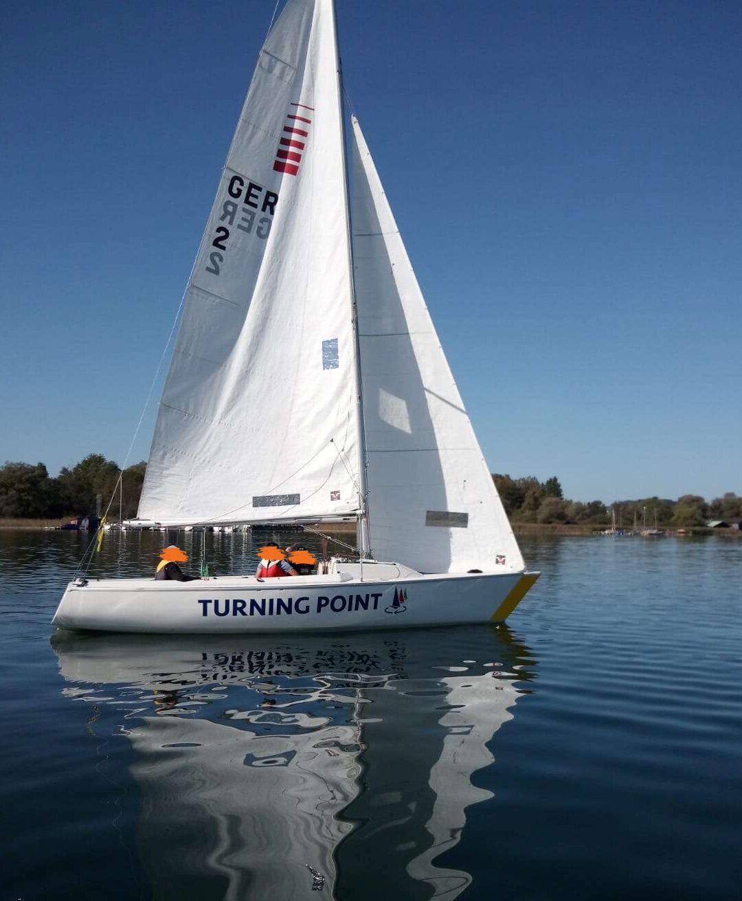 Eine weiße Segeljacht namens Turning Point mit den drei Mitgliedern an Bord schwimmt auf dem blauen Wasser. Die Segel sind angehoben und der Himmel ist blau und klar. Die Jacht spiegelt sich auf dem Wasser. In der Ferne gibt es Bäume und einige weitere Boote.