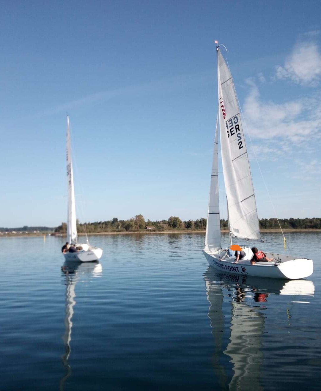 Zwei weiße Segelboote schwimmen auf dem Wasser. Auf den Segeln stehen die Buchstaben G, E, R und S. Die Besatzungsmitglieder sind nicht deutlich zu erkennen. Im Hintergrund sind Bäume und eine blaue Himmel zu sehen.