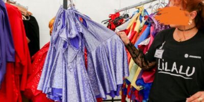 Eine Frau hält ein lilafarbenes Cape in der Hand. Hinter ihr sind mehrere Kleiderstangen zu sehen, an denen weitere bunte Kinderkostüme hängen.