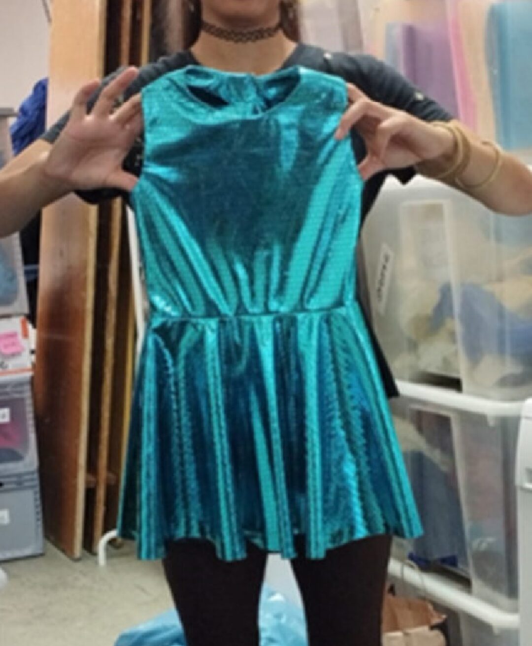 Eine Person hält ein türkisfarbenes Kinderkleid vor sich. Hinter der Person sind mehrere Kisten mit Kinderkleidungen drinnen zu sehen.