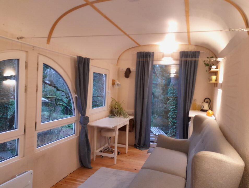 Ein gemütlicher Raum mit vielen Fenstern, die einen Blick in den Garten offenbaren. Der Raum ist hell, mit dunklen Vorhängen an den Fenstern, einem Holzboden, einem hellen Teppich und einer grauen Couch. Es wirkt gemütlich und entspannend.