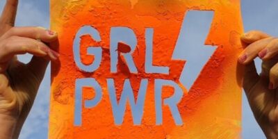 Eine Person hält ein buntes Blatt Ppaier mit der Aufschrift „Girl PWR“ in der Hand. HinTer ihr sieht man einen blauen Himmel.