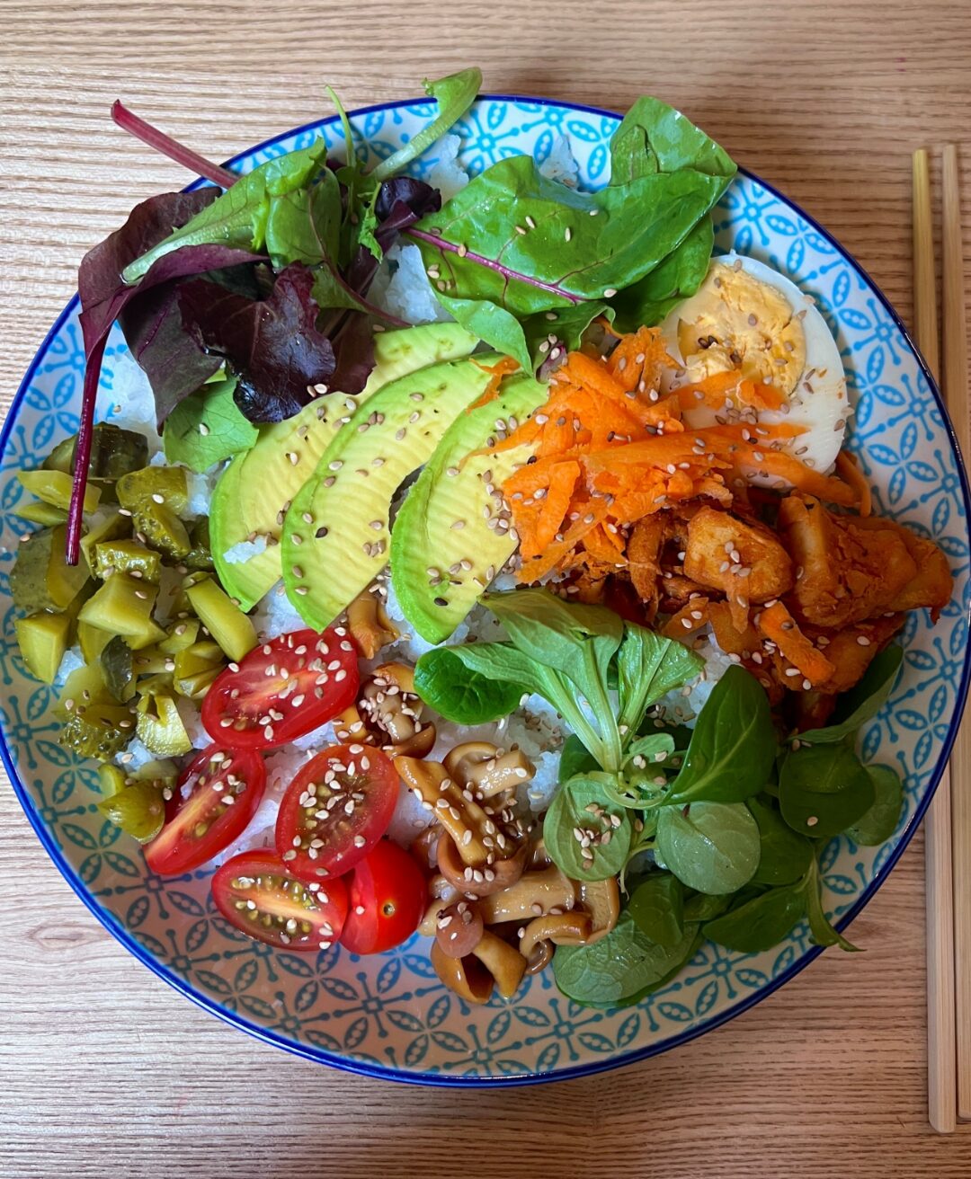 Eine Schüssel mit einer Bowl mit Reis und darauf einiges an Gemüse, wie unter anderem Avocado, Ei, Rucola, Salat, Jalapenos, Tomaten, Champingnos. Neben der Schüssel liegen Holzstäbchen.