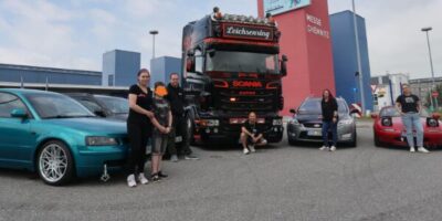Eine Gruppe von Menschen steht vor einem großen schwarzen LKW mit der Aufschrift "SCANIA" und "Leichsernring". Sie sind umgeben von mehreren Autos und einem Gebäude im Hintergrund.