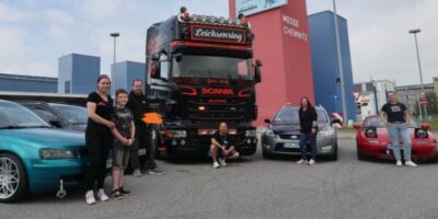 Eine Gruppe von Menschen steht vor einem großen schwarzen LKW mit der Aufschrift "SCANIA" und "Leichsernring". Sie sind umgeben von mehreren Autos und einem Gebäude im Hintergrund.
