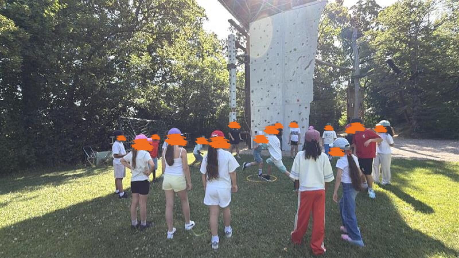 Eine Gruppe von Kindern steht vor einer Kletterwand und scheint sich auf eine Aktivität vorzubereiten. Einige Kinder tragen Rucksäcke und Schuhe, was darauf hindeutet, dass sie für eine Klettertour ausgerüstet sind. Die Kletterwand ist mit Griffen und Haltepunkten ausgestattet, die den Kindern beim Klettern helfen. Im Hintergrund sind Bäume und ein Zaun zu sehen, die die Umgebung abgrenzen.