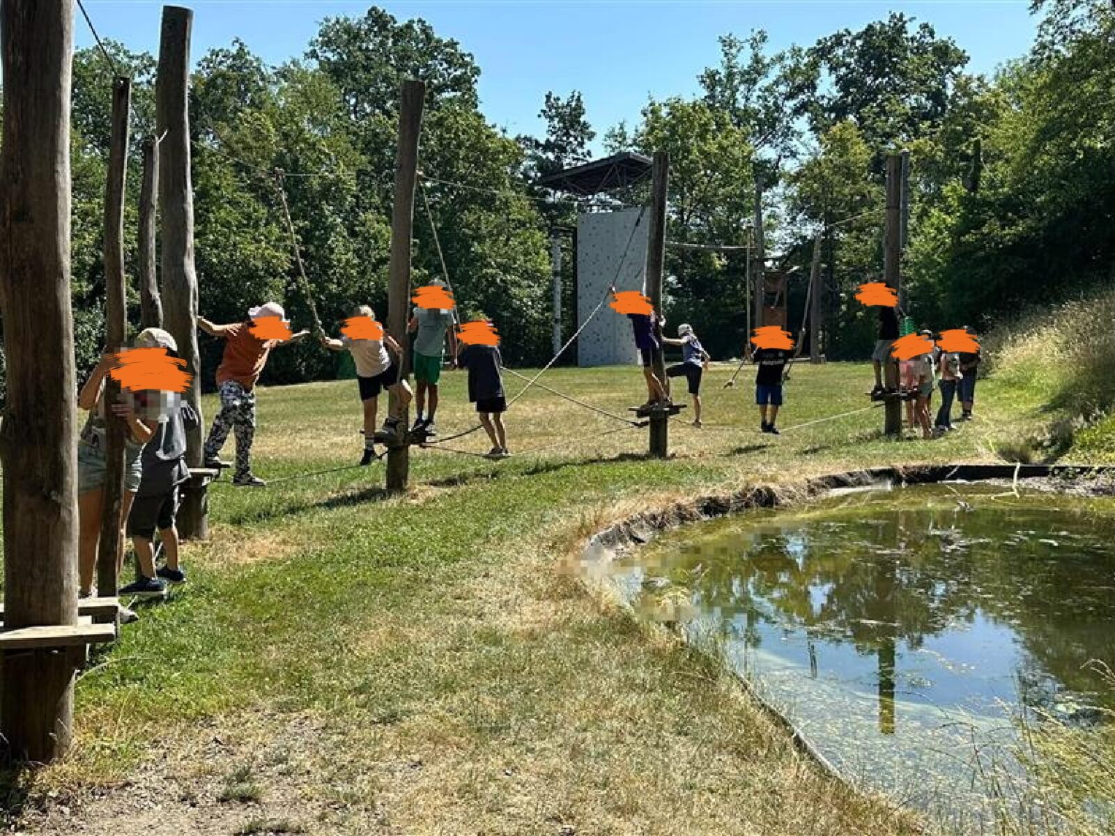 Eine Gruppe von Kindern, deren Gesichter unkenntlich gemacht wurden, nimmt auf einem Spielplatz an einer Seilbahn-Aktivität teil. Die Kinder stehen auf Plattformen, die durch Seile verbunden sind, und scheinen sich auf die Übung vorzubereiten. Im Hintergrund ist ein Teich zu sehen, der von Gras und Bäumen umgeben ist, was auf eine natürliche und entspannende Umgebung hinweist.